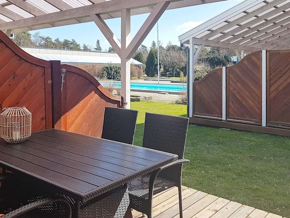 A wooden deck with a table and chairs, a pergola overhead, and a view of a swimming pool and lawn.