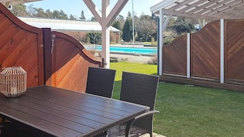 En udendørs terrasse med et træbord og stole, en pergola over hovedet og udsigt til en swimmingpool i baggrunden.