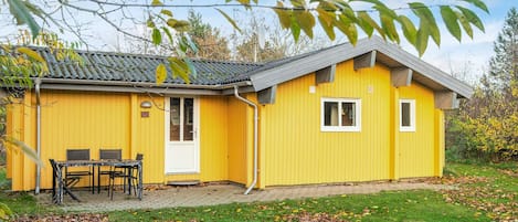 Un edificio amarillo con techo de tejas, una puerta y una ventana. Hay una mesa y sillas afuera.