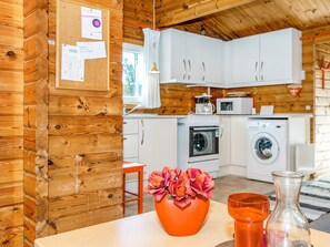A cabin interior with a wooden wall, a bulletin board, a table with a vase of flowers, and a kitchen area with appliances. - Reposeful Holiday Home in Skjern near Sea (Skjern)