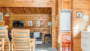 Interior acolhedor de uma cabana, paredes de madeira, televisão, área de refeições com cadeiras, cozinha com geladeira e uma pequena mesa com chaleira.