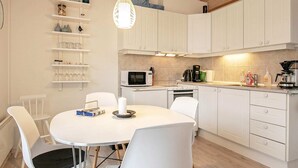 A modern kitchen with white cabinets, a round dining table, and chairs. There is a microwave, coffee maker, and a shelf with glassware. - 4 Person Holiday Home in Aeroskobing (Aeroskobing)