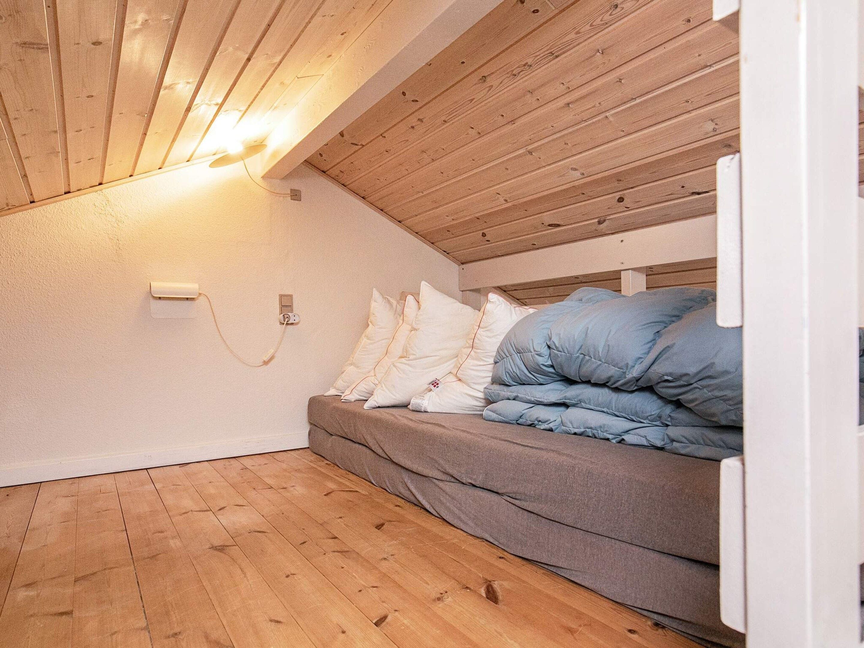 A small, cozy room with a sloped ceiling, wooden flooring, a bed with pillows, and a wall-mounted light fixture.