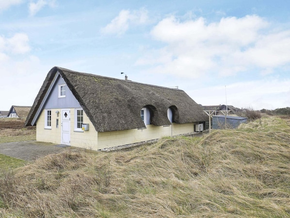 Une maison à toit de chaume, avec une seule porte et deux fenêtres, entourée d’un terrain herbeux.