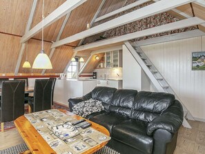 A loft-style room with a high ceiling, exposed wooden beams, a black leather sofa, a wooden coffee table with a newspaper print, and a kitchen area with white cabinets and a staircase.