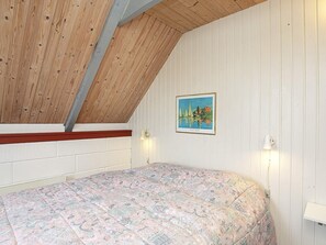 A room with a sloped ceiling, a bed with a patterned bedspread, a framed picture on the wall, and two wall-mounted lamps.