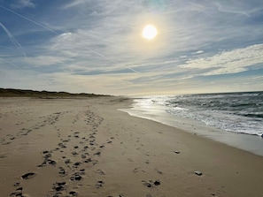 En strand med fodspor, der fører ned til vandet, en klar himmel og solen lavt på horisonten.