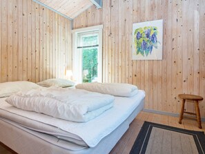 A bedroom with a wooden paneled wall, a bed with white bedding, a small stool, and a window with blinds.
