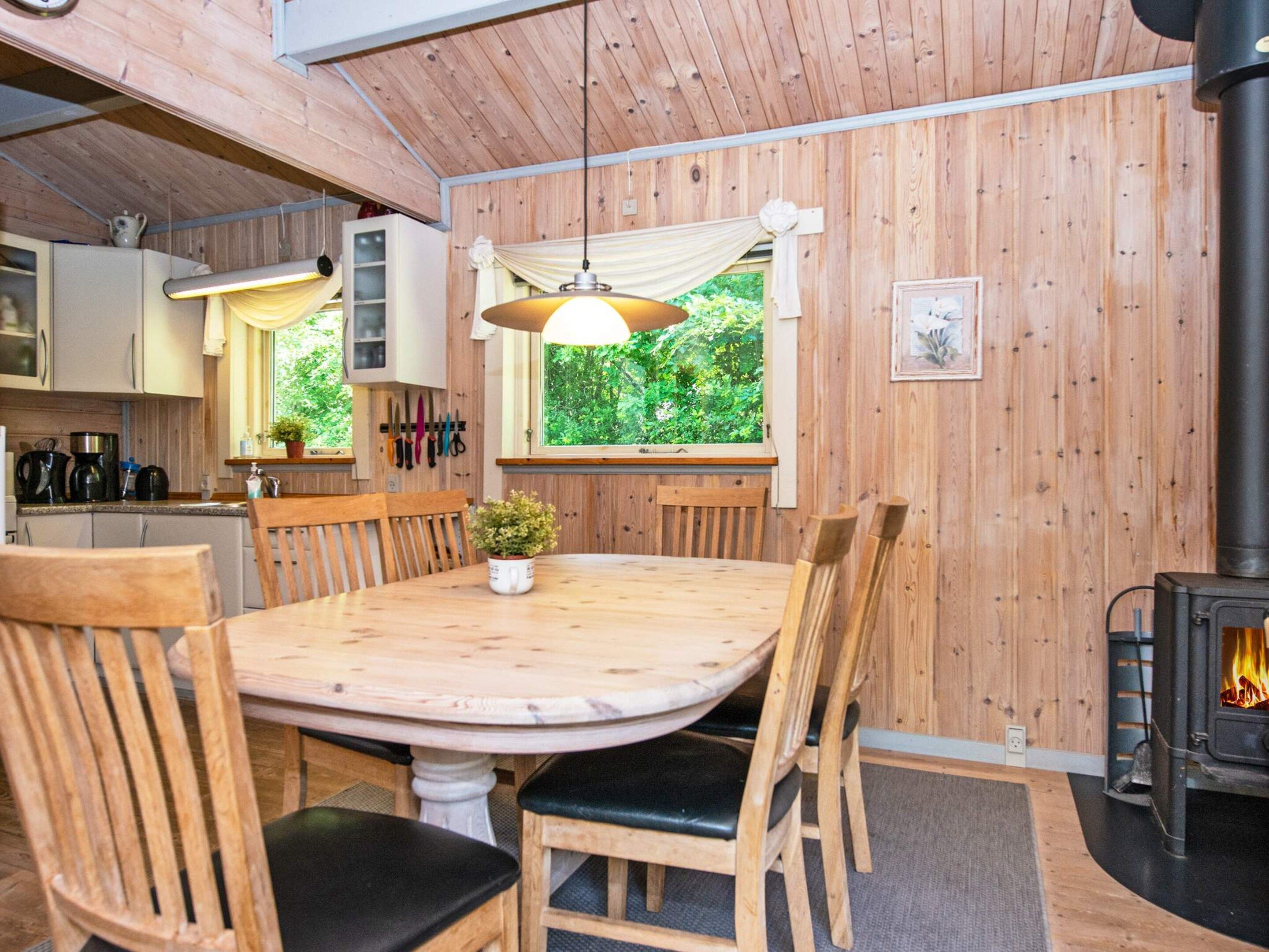A wooden interior with a dining table, chairs, a hanging light fixture, a window with a view of greenery, and a wood-burning stove.