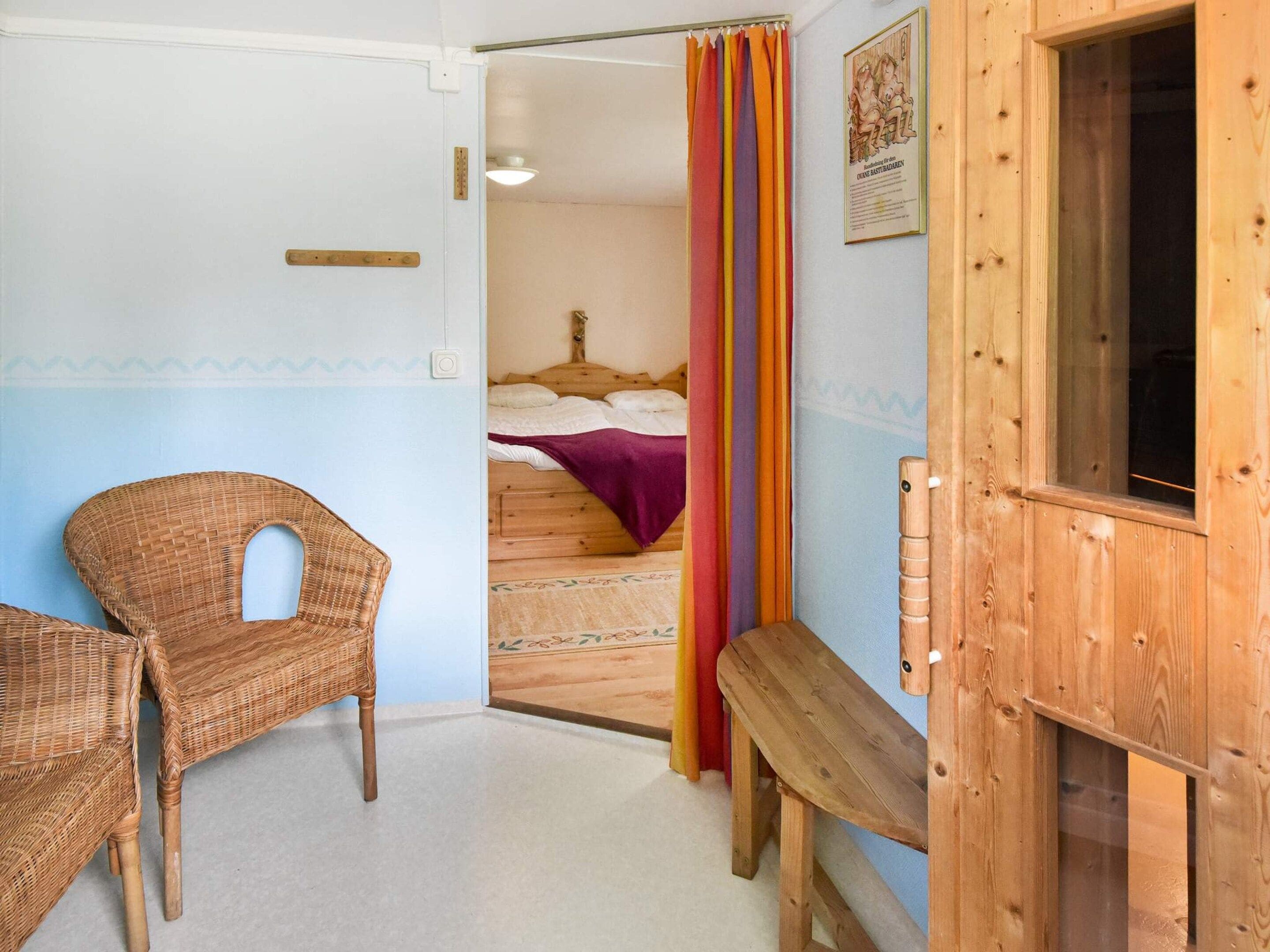 A room with a wooden bench, a wicker chair, a bed with a colorful curtain, and a wooden door.