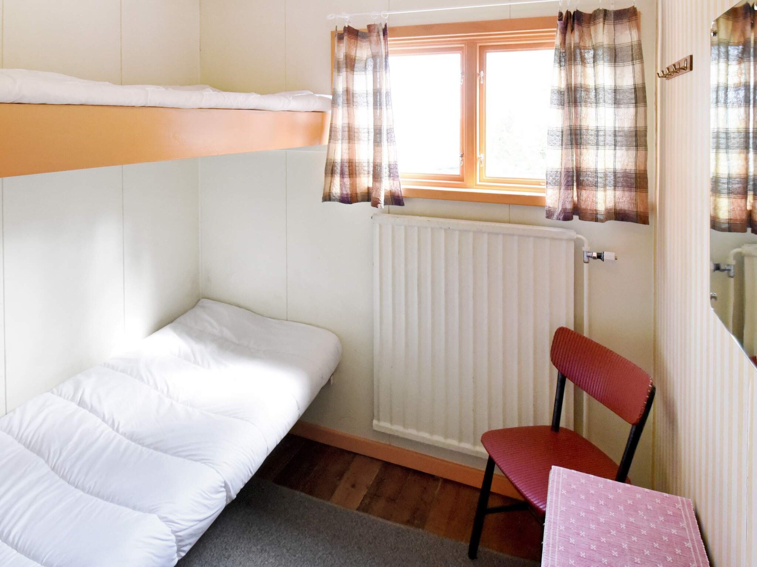 Habitación pequeña y funcional con una cama litera, una silla individual, una mesa y un ventanal con cortinas.