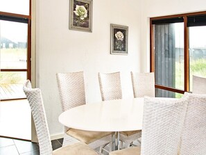 A dining area with a round table and six chairs, two framed floral paintings on the wall, and large windows with a view of greenery.