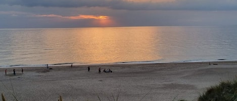 En strand med folk som promenerar och sitter, och solen som går ner över vattnet.