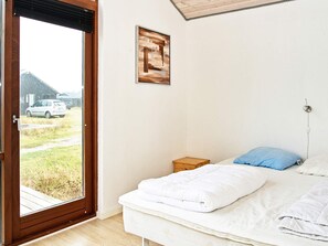 A bedroom with a bed, a small bedside table, a wall-mounted power outlet, and a window with a view of a car and buildings outside.