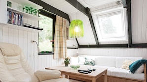 Een knus vertrek met een witte bank, een lederen fauteuil, een houten tafel en een groene hanglamp.