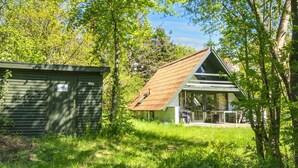 Een rustieke hut met schuin dak en een veranda, omringd door weelderige begroeiing en bomen.