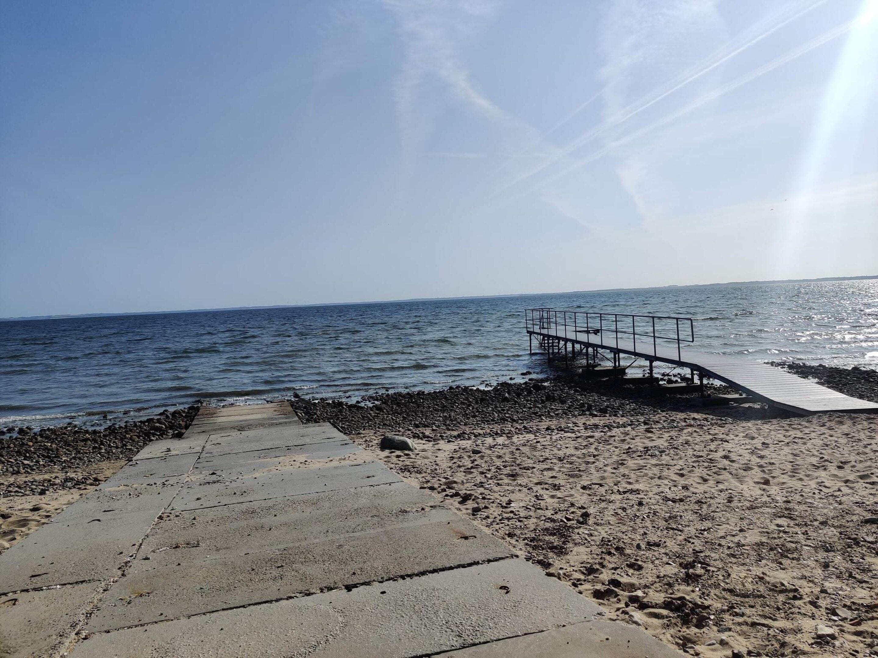 Un camino de hormigón que lleva a un muelle de madera que se adentra en el mar, con una playa rocosa al frente.
