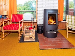 Ein gemütliches Zimmer mit Kaminofen, einem rot gestreiften Sessel, einem Korbkorb mit Holz und Blick ins Grüne.