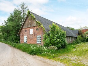 A rustic brick building with a sloped roof, surrounded by lush greenery and wildflowers. - 5 Person Holiday Home in Hojer (Højer)