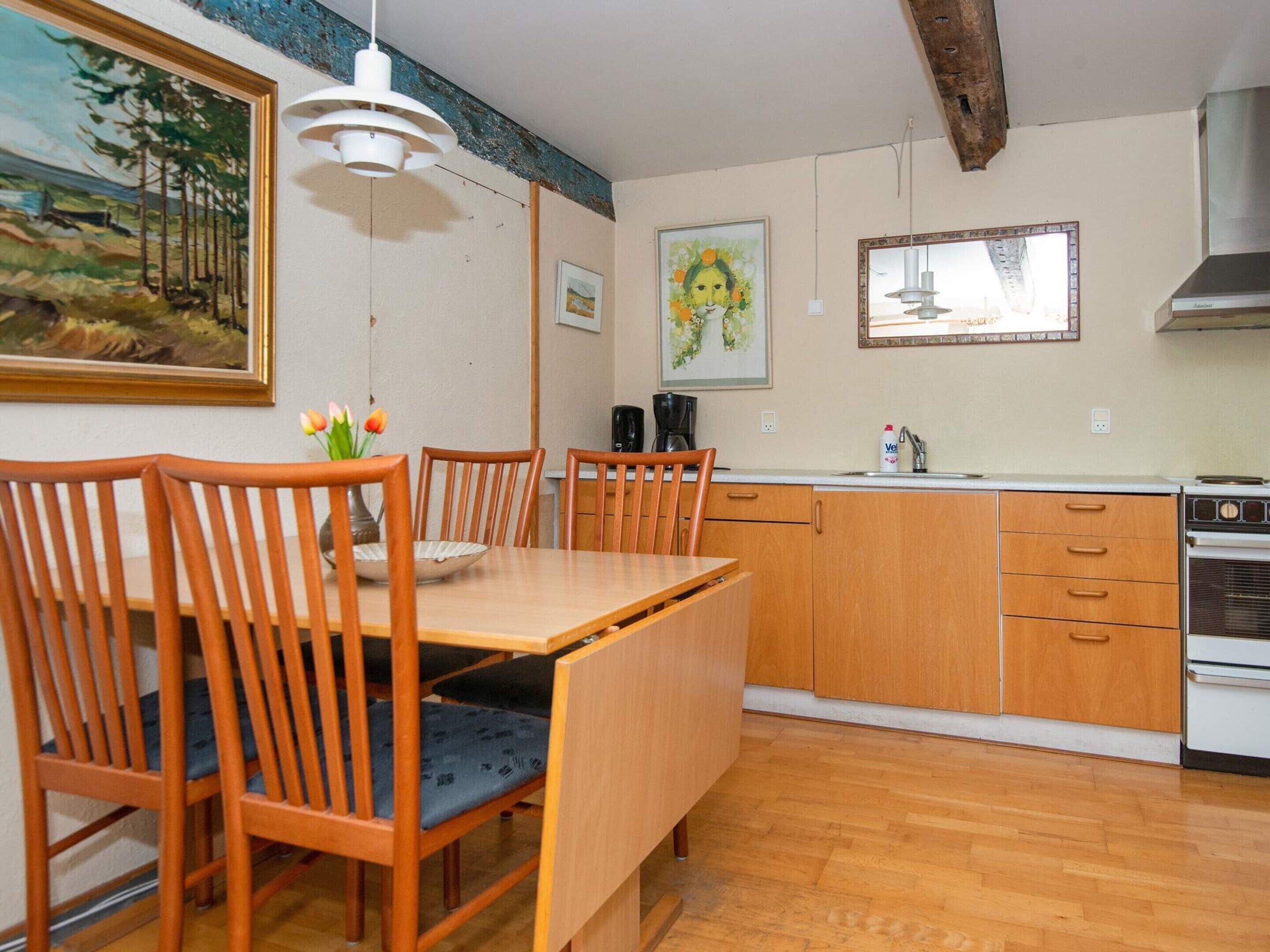 A dining area with wooden chairs and a table, a kitchen with wooden cabinets and a stove, and artwork on the walls.