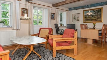Una sala de estar acogedora con una mesa de comedor de madera, un sofá y un televisor. Hay ventanas con cortinas, un radiador y un cuadro en la pared.