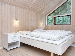 A bedroom with a wooden ceiling, a bed with a white mattress and pillows, a bedside table with drawers, and a window with a view of greenery. - 5 Star Holiday Home in Ulfborg (Ulfborg)