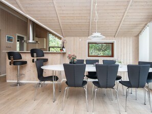 Un intérieur moderne avec un plafond en bois, une table à manger ronde entourée de chaises, un coin cuisine avec un fourneau et une fenêtre donnant sur un paysage verdoyant.