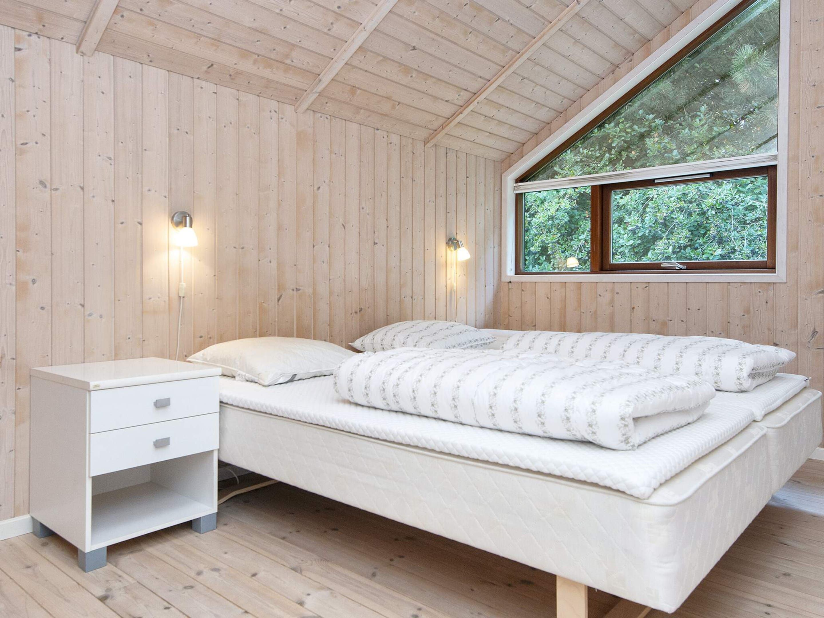 Un dormitorio con techo de madera, cama con colchón blanco y ropa de cama floral, mesita de noche con cajones y ventana con vista a follaje.