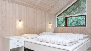 Un dormitorio con techo de madera, cama con colchón blanco y ropa de cama floral, mesita de noche con cajones y ventana con vista a follaje.