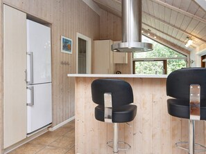 Une cuisine avec des murs en bois, une hotte de cuisine en acier inoxydable, un réfrigérateur blanc et deux tabourets de bar noirs.