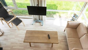 A living room with a beige sofa, a wooden coffee table with a remote control, a black chair with a woven seat, and a TV on a stand. - 6 Person Holiday Home in Sjolund-by Traum (Sjølund)