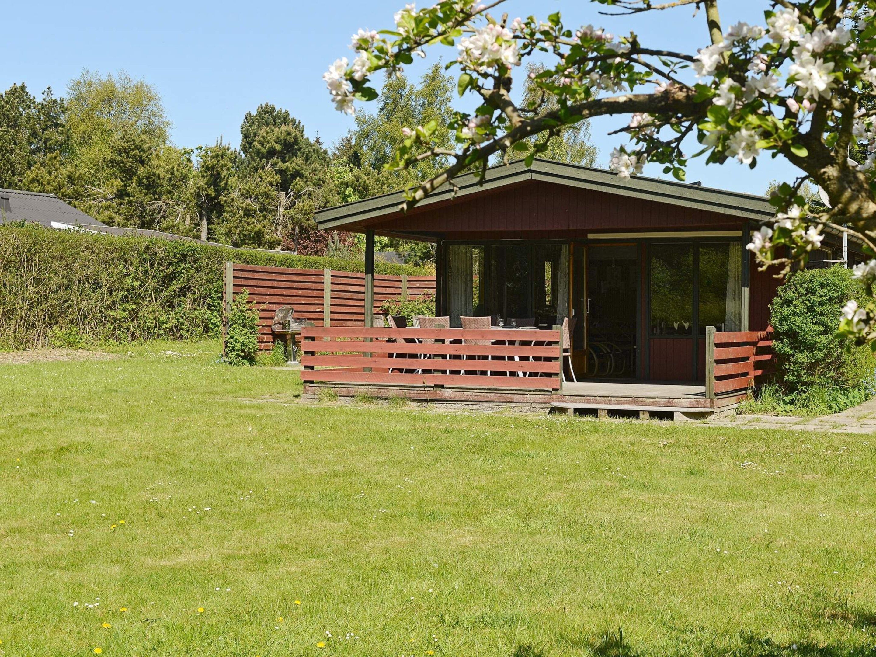 A small wooden structure with a covered porch, situated in a grassy area with trees and a hedge in the background.