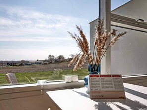 A view from a window with a vase of dried plants, a table with a glass and a menu, and a distant view of houses and a field.