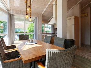 A dining area with a wooden table and wicker chairs, a kitchen in the background, and large windows with curtains.