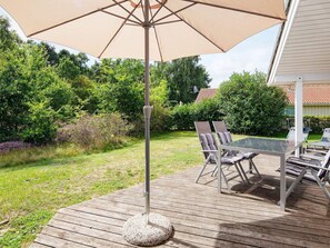 A wooden deck with a glass table and chairs, a beige umbrella, and a garden with trees and shrubs.