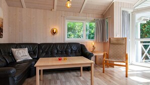 A cozy room with a black leather sofa, a wooden coffee table, and a wooden chair. There is a window with curtains, a wall-mounted lamp, and a ceiling light fixture. - Holiday Home in Groemitz (Groemitz)