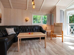 A cozy room with a black leather sofa, a wooden coffee table, and a wooden chair. There is a window with curtains, a wall-mounted lamp, and a ceiling light fixture. - Holiday Home in Groemitz (Groemitz)