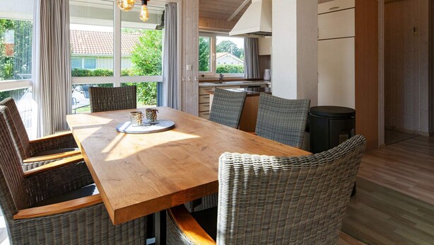 A dining area with a wooden table and wicker chairs, a kitchen in the background, and large windows with curtains. - Holiday Home in Groemitz (Groemitz)