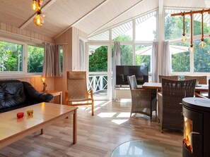 A spacious room with a wooden floor, a television, wicker furniture, and large windows offering a view of greenery.
