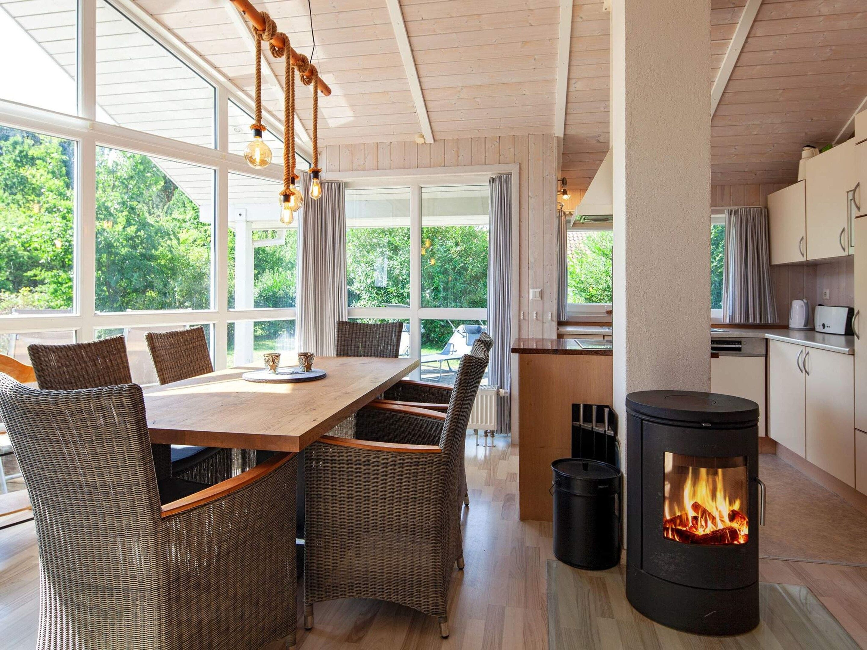 A modern kitchen with a wood-burning stove, large windows, and a dining area with wicker furniture.