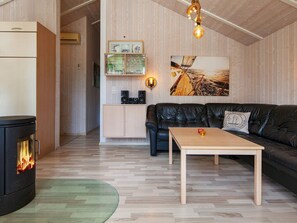 A cozy living room with a fireplace, a leather sofa, a wooden coffee table, and a kitchen area with cabinets and a hanging light fixture. - Holiday Home in Groemitz (Groemitz)