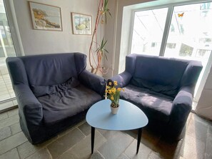 Quarto com dois sofás escuros, uma mesinha redonda com planta em vaso e uma janela com vista para edifícios.