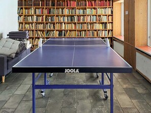A ping pong table with a blue frame and 'JOOLA' branding, positioned in a room with a bookshelf filled with books in the background.