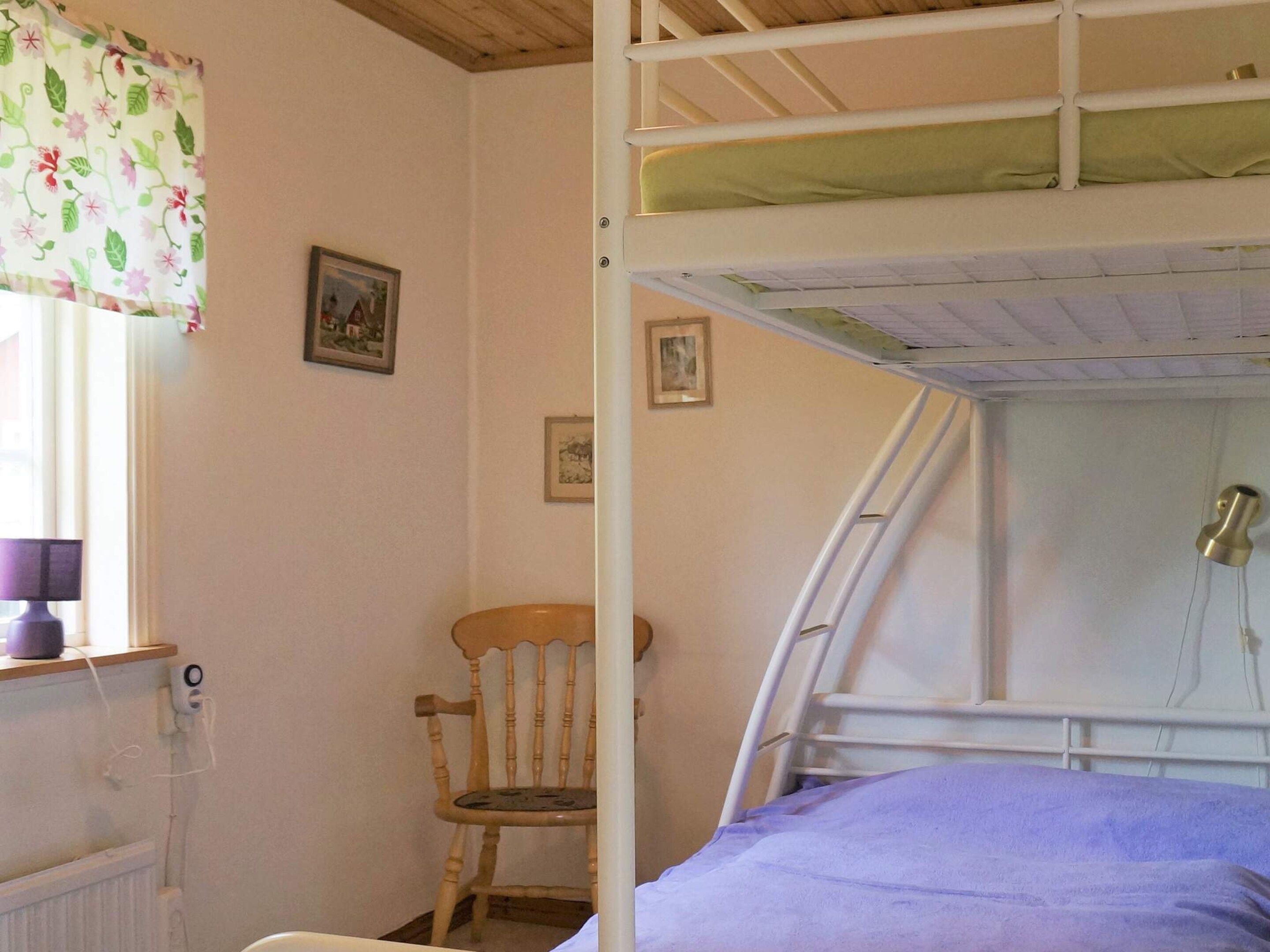Uma cama beliche com colchão verde, uma cadeira de madeira e uma janela com cortina florida.