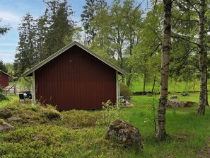 A red wooden building with a gabled roof, surrounded by a grassy area with trees and rocks. - 6 Person Holiday Home in Asarp (Åsarp)