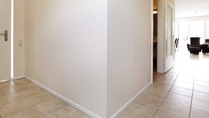A hallway with a door on the left, a light fixture on the ceiling, and a glimpse of a seating area with chairs and a table in the background. - Holiday Home in Grossenbrode (Grossenbrode)