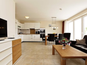 A modern kitchen with white cabinets, a dining area with a wooden table and chairs, and a living room with a sofa and a coffee table. - Holiday Home in Grossenbrode (Grossenbrode)