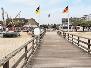 A wooden boardwalk leading to a beach area with flags, buildings, and a Ferris wheel in the background. - Holiday Home in Grossenbrode (Grossenbrode)