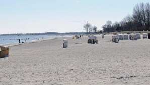 A sandy beach with a calm sea, a few people, and some structures that appear to be storage or utility units. - Holiday Home in Grossenbrode (Grossenbrode)