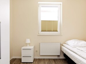 A bedroom with a bed, a nightstand with a lamp, a radiator, and a window with blinds. - Holiday Home in Grossenbrode (Grossenbrode)